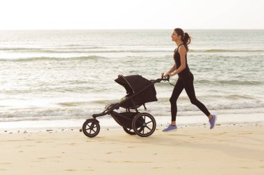Young mom is running with stroller on the beach. clipart