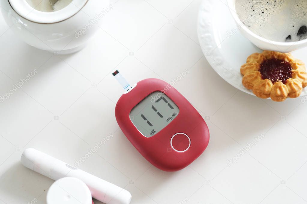 Glucometer showing high level on glucose lying next to cup of coffee and sweet cookies.