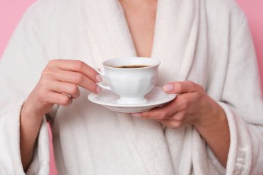 Kahve havası. Beyaz banyolu kadın elinde bir fincan kahve tutuyor. Pembe arka planda stüdyo çekimi.