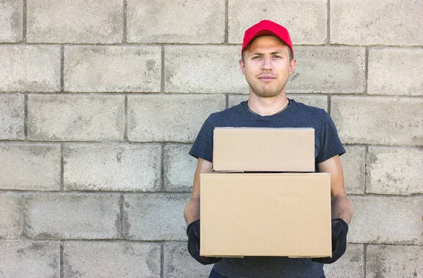 a young man delivery man stands against the background of a gray stone wall, holds two cardboard boxes in his hands, in a red cap and gray T-shirt, delivery, place for an inscription