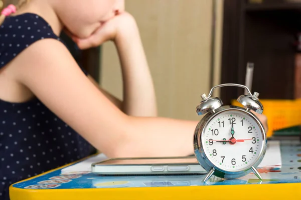 Het meisje zit aan tafel en doet haar huiswerk, de klok op de voorgrond toont negen oclock in de ochtend, tijd voor lessen, afstandsonderwijs, klok gezicht — Stockfoto