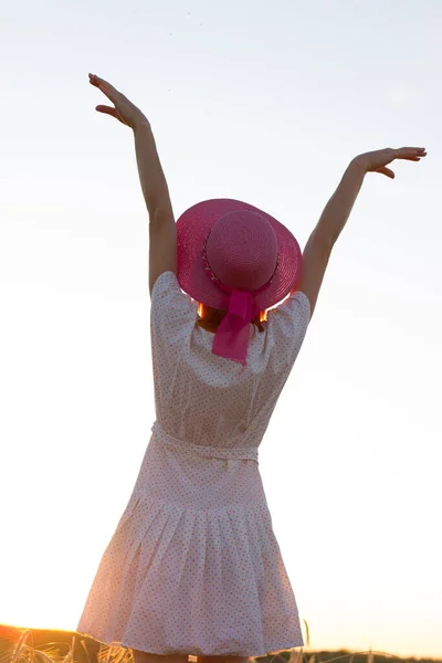 Ένα κορίτσι στέκεται με την πλάτη στον ουρανό το ηλιοβασίλεμα, τα χέρια σηκωμένα, μια γυναίκα με ροζ καπέλο και λευκό φόρεμα. — Φωτογραφία Αρχείου