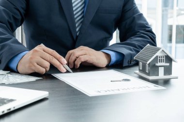 Emlakçının eli, elinde bir kalem ve ev sigortası sözleşmesi belgeleriyle birlikte müşterilerine sunacağı ev modelleri ve anahtarlarla ilgili bir anlaşma hazırlıyor..