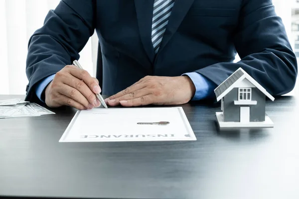 The hand of the real estate agent, holding a pen and drafting an agreement on the home insurance contract documents, along with samples of house models and keys to present to his clients.