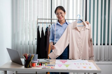 Tekstil aksesuarları ve girişimci tasarımcı çizimleriyle genç ve güzel Asyalı moda terzisi stüdyoda yaratıcı fikirler için dizüstü bilgisayarı olan parlak renklerle dolu..