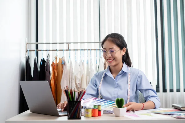 Tekstil aksesuarları ve girişimci tasarımcı çizimleriyle genç ve güzel Asyalı moda terzisi stüdyoda yaratıcı fikirler için dizüstü bilgisayarı olan parlak renklerle dolu..