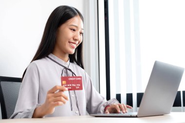 Asyalı iş kadınının elinde bir kredi kartı var ve ofiste online alışveriş ve internet ödemeleri için dizüstü bilgisayar kullanıyor..