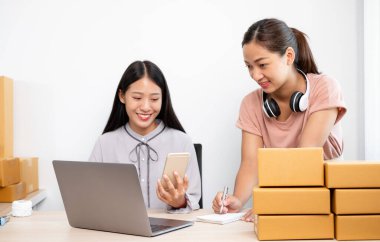 Asyalı kadın işi. Masasında bir sürü paket kutusu olan iki kişi mutlu bir online satış işi, bilgisayarınızı kullanın, müşterilerden emir alın, notlar alın ve posta yoluyla teslimat için düzenlemeler yapın..
