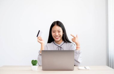 Asyalı iş kadınının elinde bir kredi kartı var ve ofiste online alışveriş ve internet ödemeleri için dizüstü bilgisayar kullanıyor..