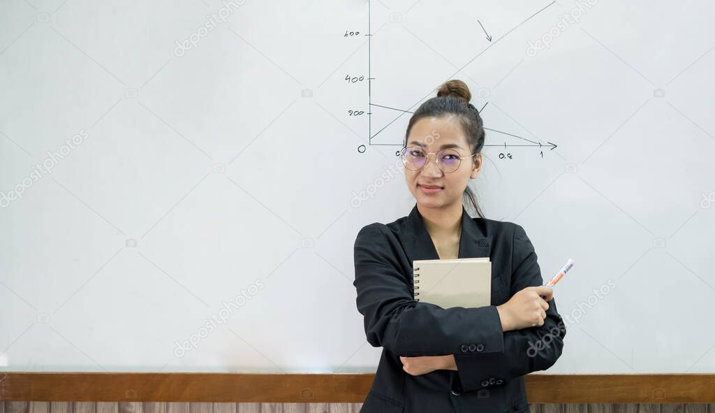 An Asian female teacher in a classroom near the whiteboard is writing and teaching a lesson with a special lecture in a class column. Math teacher explains arithmetic sums to university students.