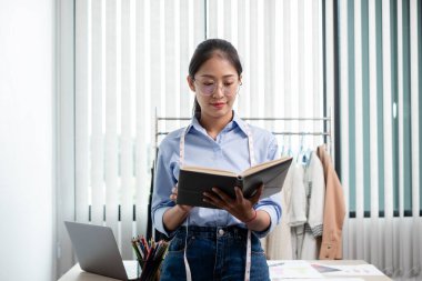 Tekstil aksesuarları ve girişimci tasarımcı çizimleriyle genç ve güzel Asyalı moda terzisi stüdyoda yaratıcı fikirler için dizüstü bilgisayarı olan parlak renklerle dolu..