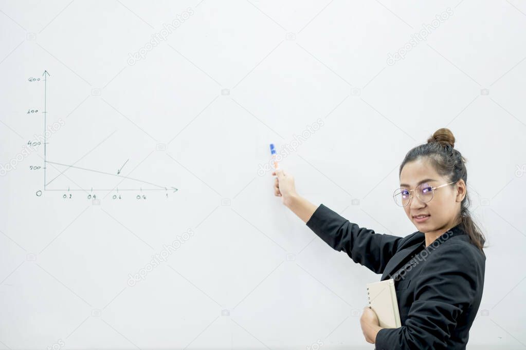An Asian female teacher in a classroom near the whiteboard is writing and teaching a lesson with a special lecture in a class column. Math teacher explains arithmetic sums to university students.