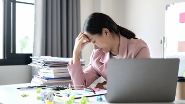 Çalışan Asyalı kadınlar stresli, işten yorgun, ofiste çalışırken çok çalışmaktan migren ağrısı çekiyor..
