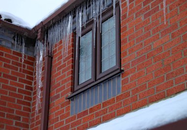Uzun buz sarkıtları bir evin oluk asmak. Çatı karla kaplı ve hala kar yağıyor.