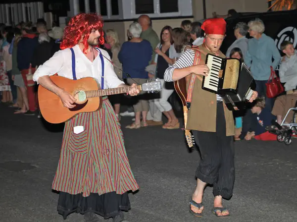 Sidmouth, Devon, England-10 augusti 2012: två artister i fancy dress spela en gitarr och en dragspel i nattetid stängning procession av folk veckan. — Stockfoto