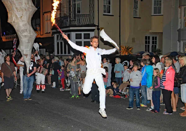 Sidmouth, Devon, England-10 augusti 2012: en mycket energisk ung man klädd i vitt och håller en trasa och en flammande fackla, dansar längs i nattetid stängning procession av folk vecka. — Stockfoto