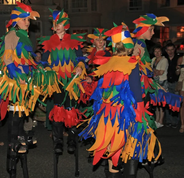Sidmouth, Devon, England-10 augusti 2012: barn utklädda till färgglada papegojor och gå på styltor delta i natten stängning procession av folk vecka. — Stockfoto