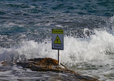 Denizdeki kayaların üzerinde kaygan bir yüzeye dair açık bir uyarı işareti var.