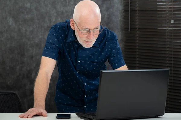 Hombre mayor con barba y camisa azul en la oficina usando el ordenador portátil —  Fotos de Stock