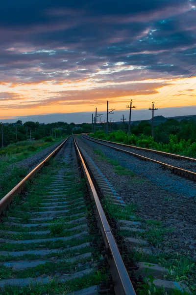 Railroad against the backdrop of sky and sunset clouds with green grass in the foreground. Beautiful sunset with orange sunbeams on a background of blue clouds. Leaving road concept. — Stock Photo, Image