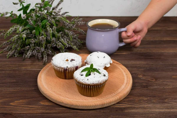 A childs hand holds a cup of tea and mint near the board with muffins with black currant, on a background of flowers and mint leaves on a wooden background — Stock Photo, Image