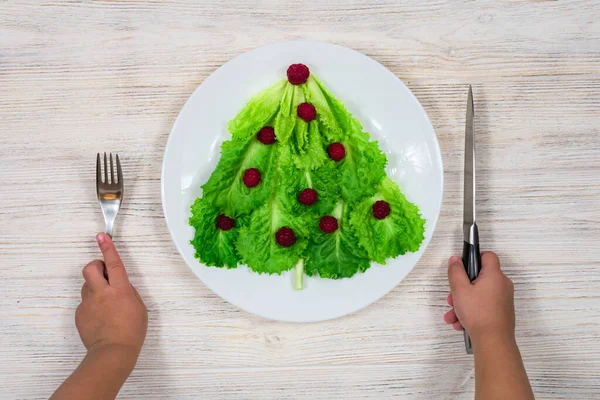 A árvore de Natal é forrada com folhas de alface verde, decoradas com framboesas vermelhas em um prato branco. As mãos das crianças estão segurando um garfo e uma faca. Comida para o Ano Novo. — Fotografia de Stock