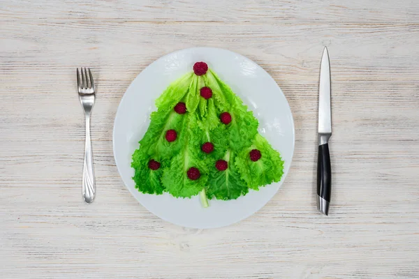 A árvore de Natal é forrada com folhas de alface verde, decoradas com framboesas vermelhas em uma placa branca com um garfo e faca. Comida para o Ano Novo. Decoração de mesa. Vista de cima. Lugar para um — Fotografia de Stock