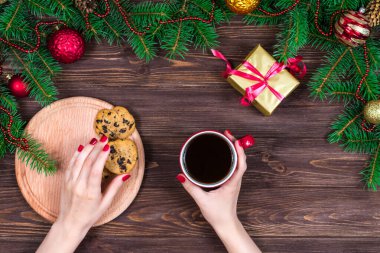 Ahşap bir masanın üzerinde bir fincan çay ve kurabiyeyle Noel ağacı ve süslemeleri olan kadın elleri.