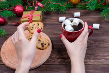 Ahşap bir masanın üzerinde bir fincan çay ve kurabiyeyle Noel ağacı ve süslemeleri olan kadın elleri.