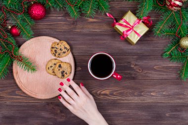 Ahşap bir masada bir fincan çay ve kurabiyeyle Noel ağacı ve süslemeleri olan bir kadın eli.