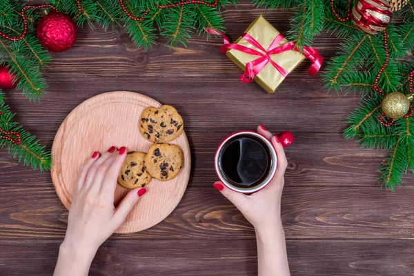 Ahşap bir masanın üzerinde bir fincan çay ve kurabiyeyle Noel ağacı ve süslemeleri olan kadın elleri.