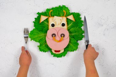 2021 'in yenilebilir boğa sembollü, yeşil marul yapraklarının üzerinde kızarmış ekmek ve sosisten yapılmış komik bir sandviç. Çocukların elleri çatal ve bıçak tutuyor. Çocuklar için kahvaltı fikri. Yeni yıl, Noel yemeği.