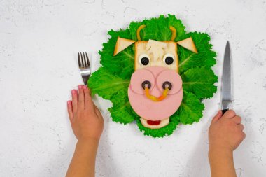 2021 'in yenilebilir boğa sembollü, yeşil marul yapraklarının üzerinde kızarmış ekmek ve sosisten yapılmış komik bir sandviç. Çocukların elleri çatal ve bıçak tutuyor. Çocuklar için kahvaltı fikri. Yeni yıl, Noel yemeği.