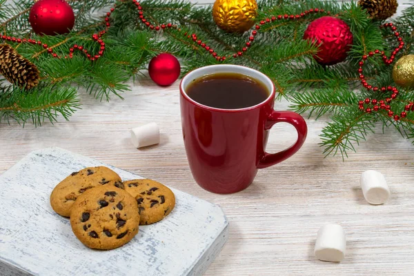 Czerwony kubek z herbatą, kawa z ciasteczkami i piankami na białym drewnianym stole na tle choinki noworocznej z dekoracjami świątecznymi. Przytulność i koncepcja Nowego Roku. — Zdjęcie stockowe
