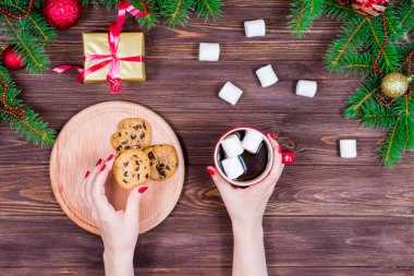 Ahşap bir masanın üzerinde bir fincan çay ve kurabiyeyle Noel ağacı ve süslemeleri olan kadın elleri.