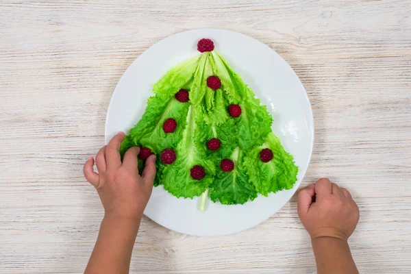 Choinka wyłożona jest zielonymi liśćmi sałaty, ozdobionymi czerwonymi malinami na białym talerzu. Dziecko zaraz zje sałatkę z choinki. Jedzenie na Nowy Rok. Dekoracja stołu. — Zdjęcie stockowe