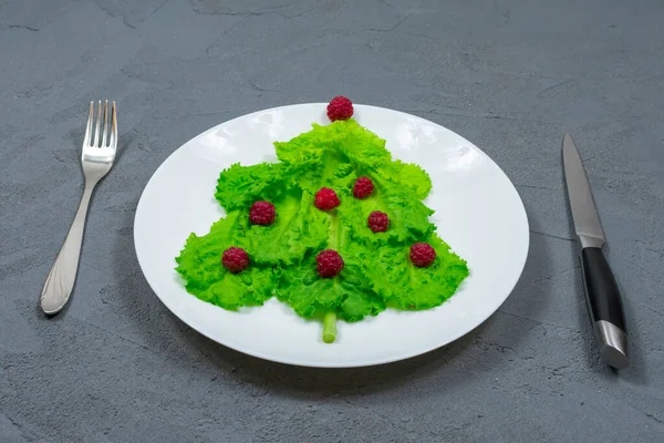 Choinka wyłożona jest zielonymi liśćmi sałaty, ozdobionymi czerwonymi malinami na białym talerzu widelcem i nożem. Jedzenie na Nowy Rok. Dekoracja stołu. Widok z góry. — Zdjęcie stockowe