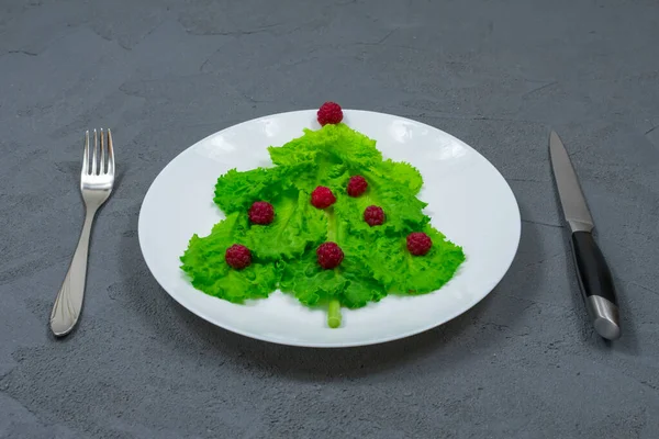 A árvore de Natal é forrada com folhas de alface verde, decoradas com framboesas vermelhas em uma placa branca com um garfo e faca. Comida para o Ano Novo. Decoração de mesa. Vista de cima. — Fotografia de Stock