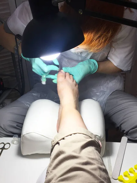 The master in the beauty salon does a professional pedicure to a young woman. Female foot in the hands of the master in the light of a lamp close-up. Tool for professional pedicure on a white table.