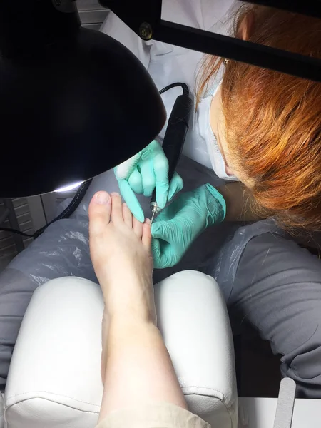 The master in the beauty salon does a professional pedicure to a young woman. Female foot in the hands of the master in the light of a lamp close-up. Tool for professional pedicure on a white table.