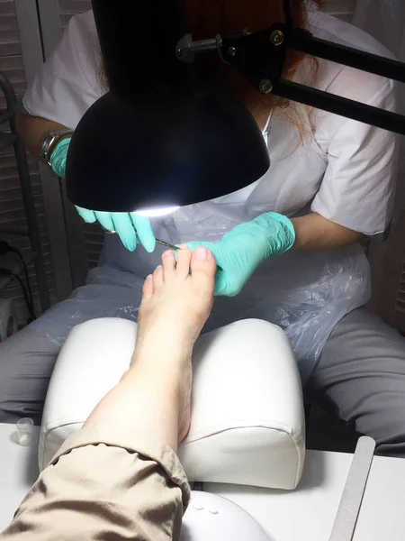 The master in the beauty salon does a professional pedicure to a young woman. Female foot in the hands of the master in the light of a lamp close-up. Tool for professional pedicure on a white table.