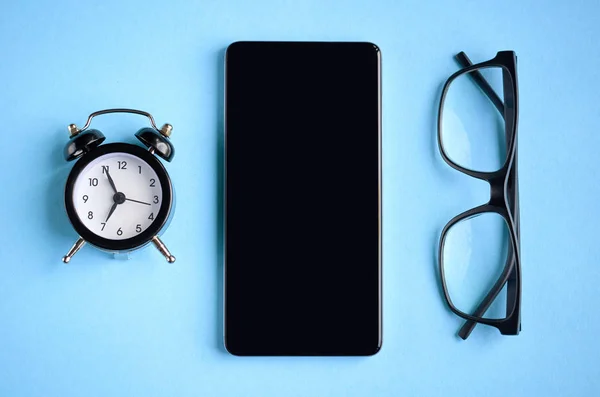 Óculos pretos, despertador e celular na composição de fundo azul . — Fotografia de Stock