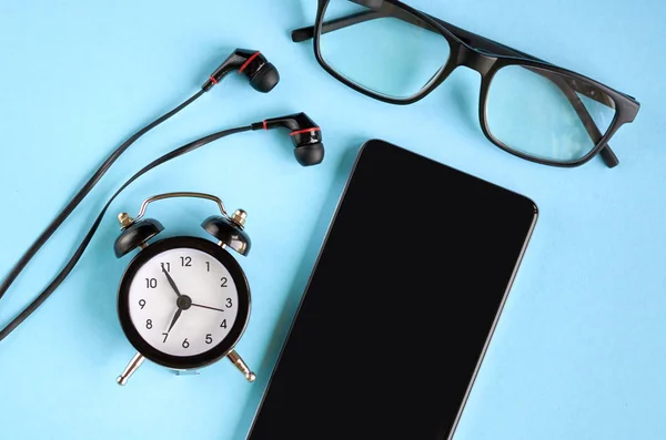 Black glasses, cellphone, alarm clock and headphones on blue background composition.
