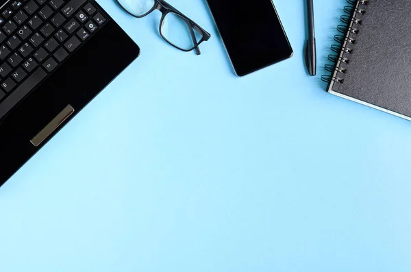 Gafas negras, teléfono celular, portátil y teclado portátil en la composición de fondo azul . — Foto de Stock