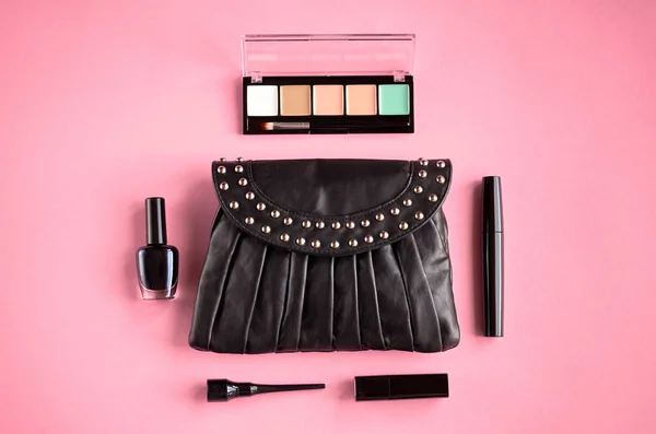 Black leather bag with nail polish, pomade, mascara and eye shadows on pink background composition. — Stock Photo, Image