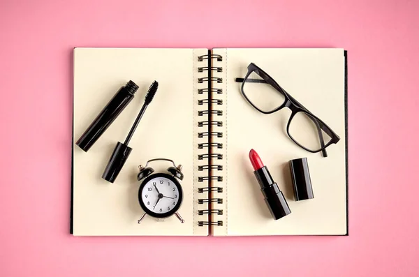 Gafas negras, despertador, bloc de notas de papel, rimel y pomada roja sobre fondo rosa composición . — Foto de Stock