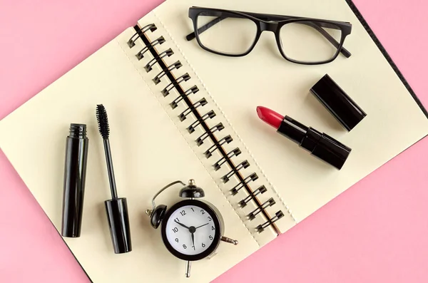 Gafas negras, despertador, bloc de notas de papel, rimel y pomada roja sobre fondo rosa composición . — Foto de Stock