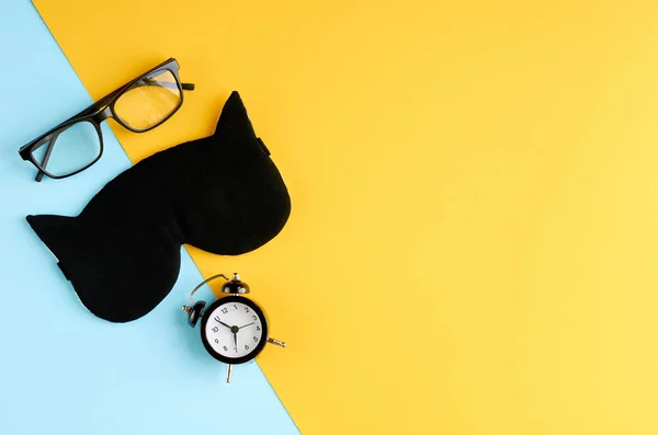 Óculos pretos, despertador e máscara de sono na composição de fundo azul e amarelo . — Fotografia de Stock