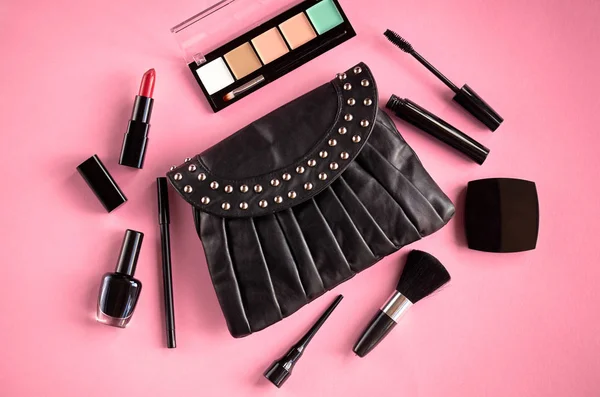 Black leather bag with nail polish, pomade, mascara and eye shadows on pink background composition. — Stock Photo, Image