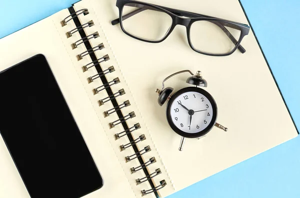 파란색 배경 구성에 검은 색 안경, 알람 시계, 종이 메모장 및 휴대 전화. — 스톡 사진